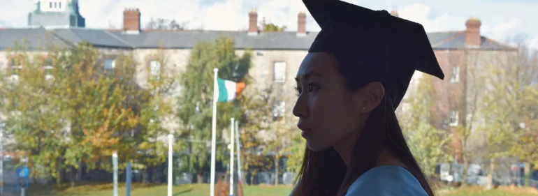 Student success at Griffith College A student in graduation cap and gown standing at a window, with the main Dublin campus in the background.